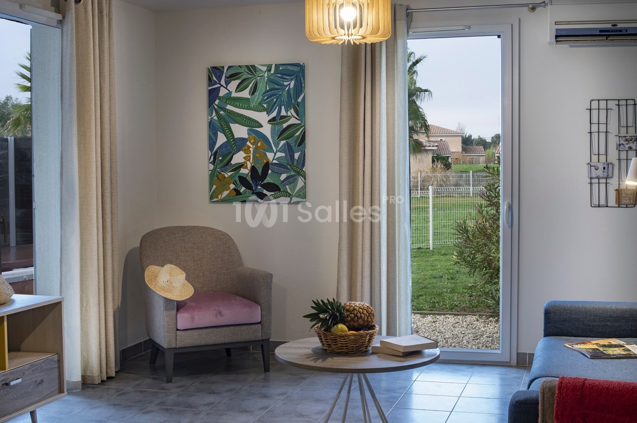 Salon lumineux avec fauteuil, table basse garnie de fruits, tableau coloré et vue sur un jardin par une porte vitrée.
