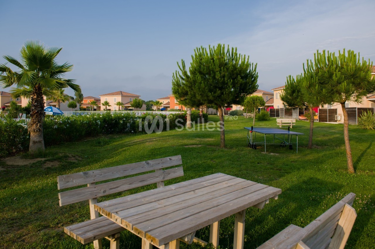Table en bois et bancs sur une pelouse, entourés d'arbres et de maisons avec une piscine visible en arrière-plan.