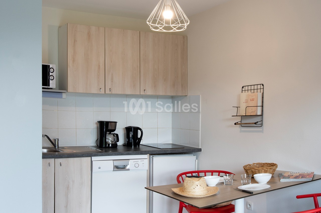 Cuisine compacte avec meubles en bois clair, table avec deux chaises rouges, cafetière, vaisselle et éclairage suspendu.