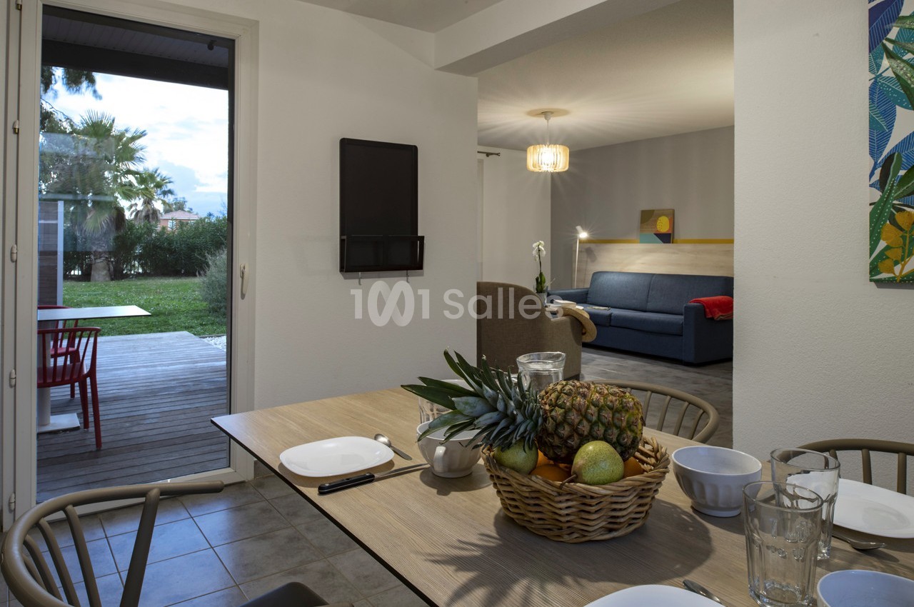 Vue d'une salle à manger avec table dressée, salon en arrière-plan et accès à une terrasse donnant sur un jardin.