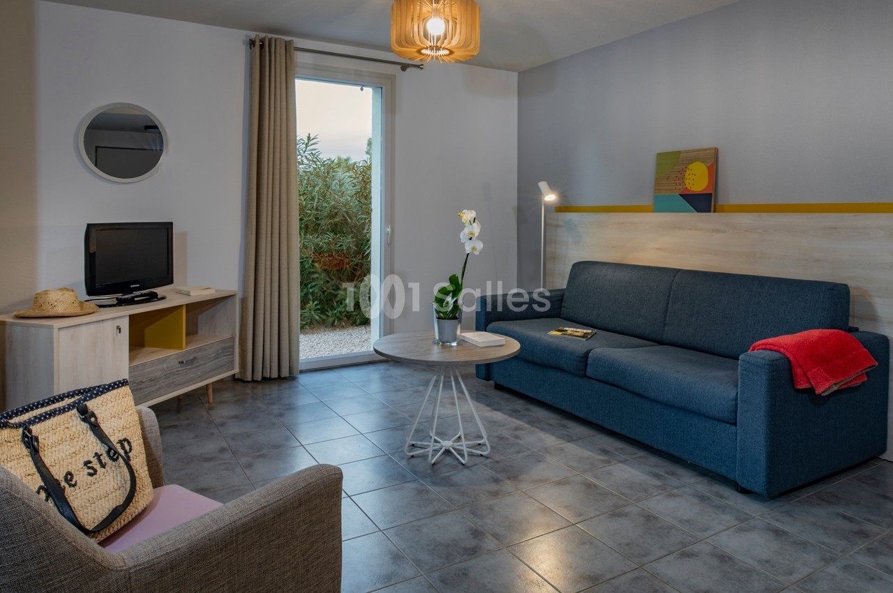 Salon moderne avec canapé bleu, table basse, fauteuil, télévision, et porte-fenêtre donnant sur un jardin.