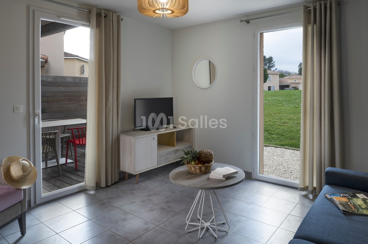 Salon lumineux avec table basse, canapé, télévision, terrasse extérieure et vue sur un jardin.