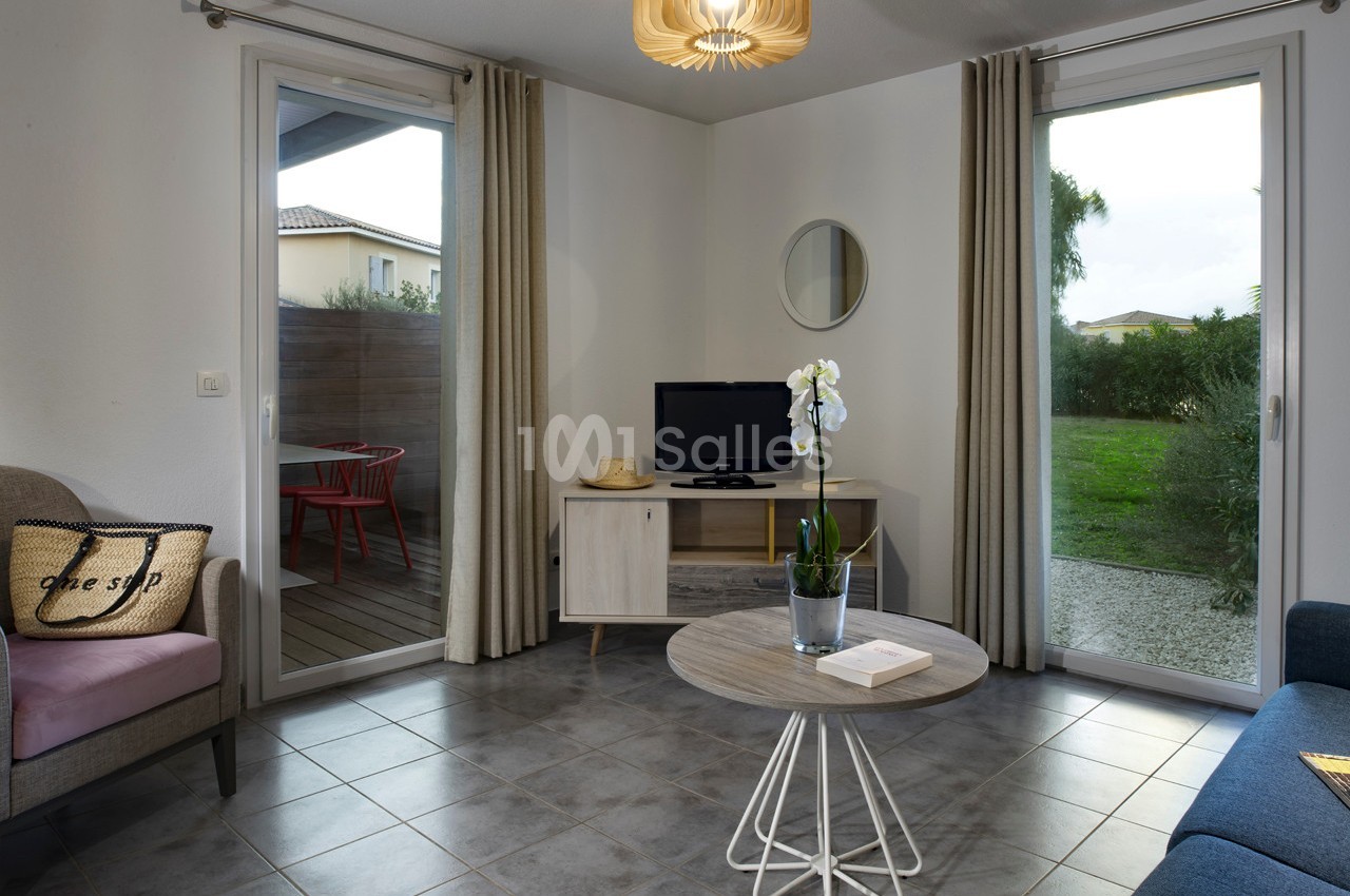 Salon lumineux avec table basse, orchidée, télévision, deux grandes fenêtres donnant sur une terrasse et un jardin.