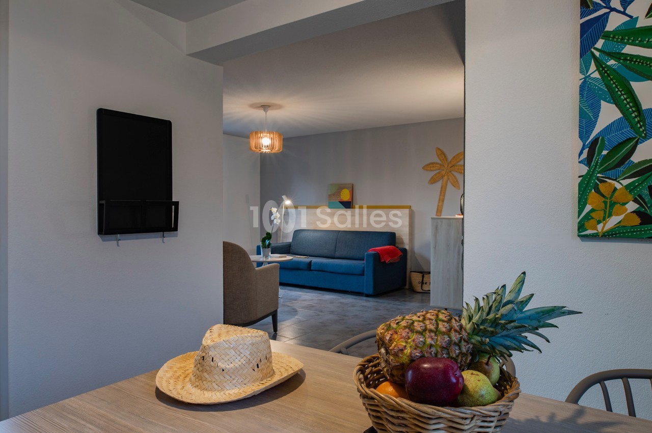 Vue d'un intérieur lumineux avec un salon en arrière-plan et une table avec un panier de fruits au premier plan.