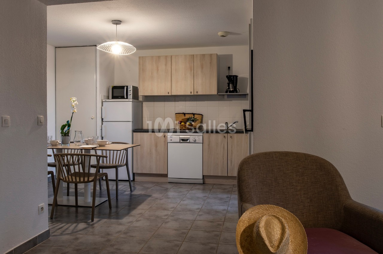 Cuisine ouverte avec table à manger, chaises en bois, électroménagers, et un fauteuil avec un chapeau posé dessus au premier…
