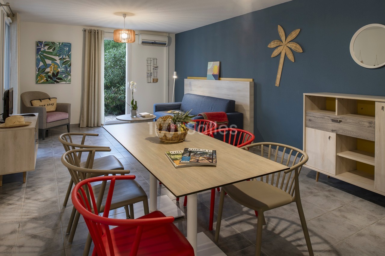 Salle à manger lumineuse avec table en bois, chaises colorées, canapé bleu, mur bleu décoré et grande baie vitrée.