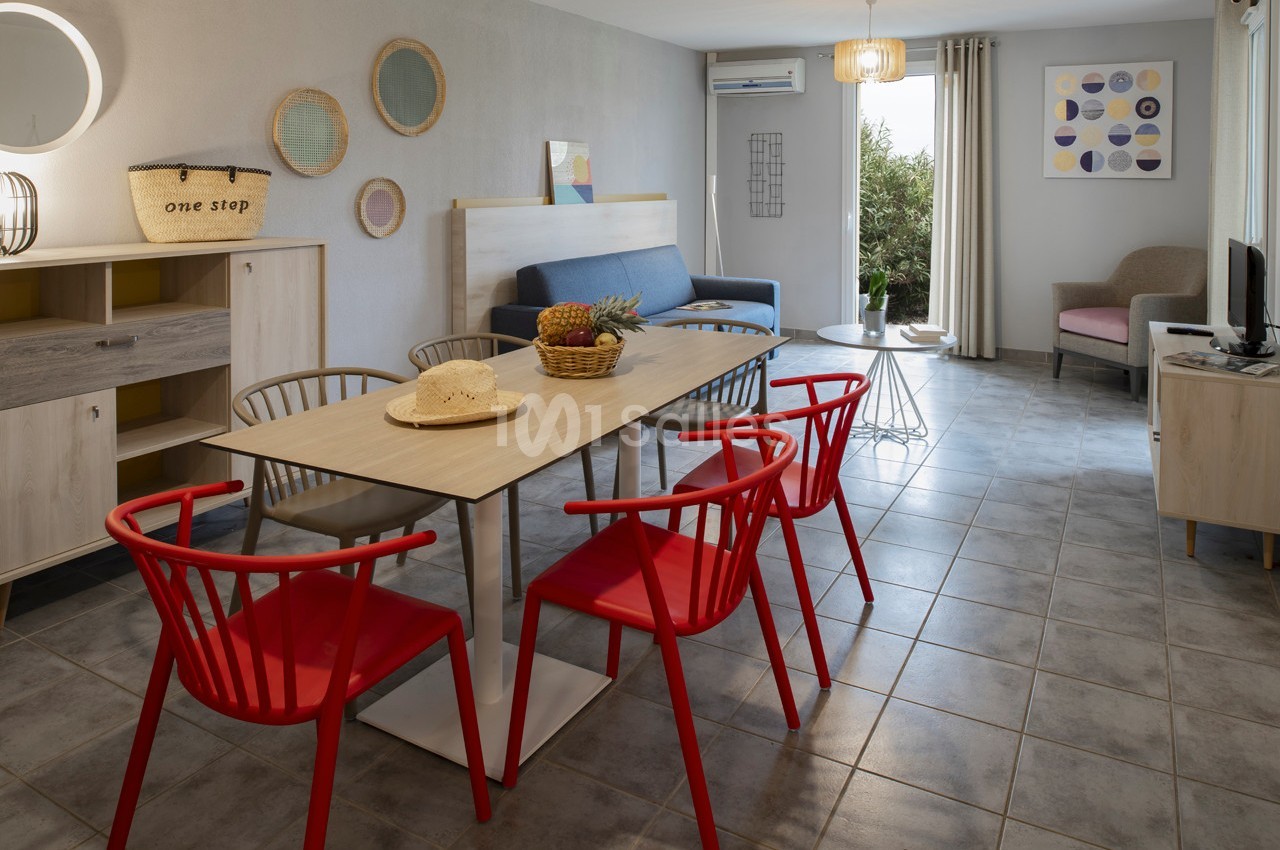 Salle à manger lumineuse avec table en bois, chaises rouges, canapé bleu et vue sur un jardin par une porte-fenêtre.
