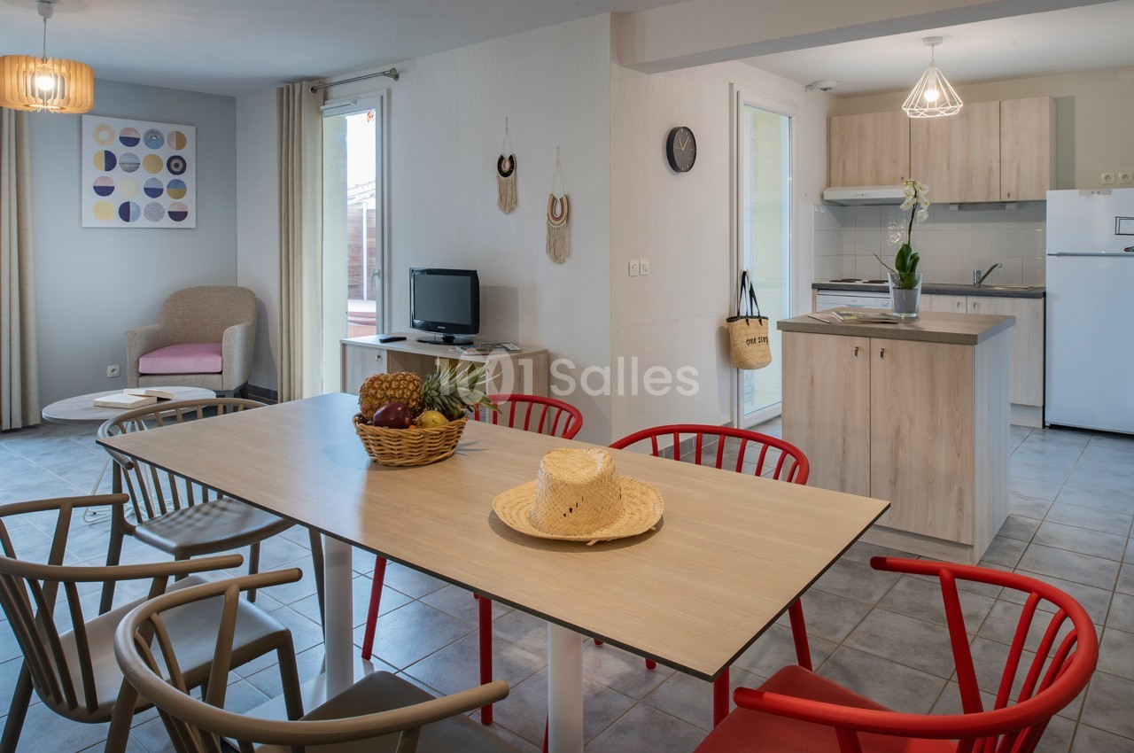 Salle à manger lumineuse avec table en bois, chaises colorées, cuisine ouverte et décoration sobre.