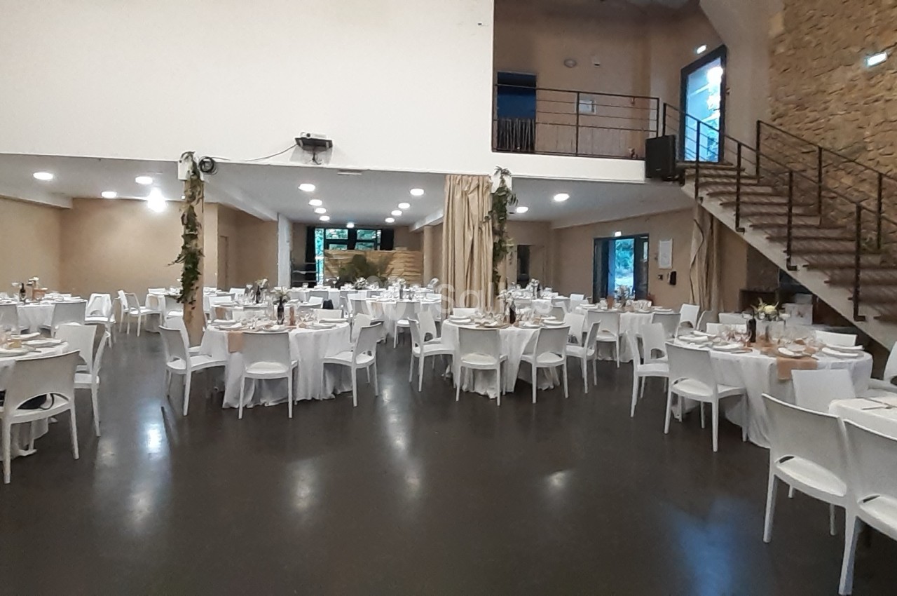 Salle de réception décorée avec des tables rondes et rectangulaires dressées pour un événement, éclairage doux.