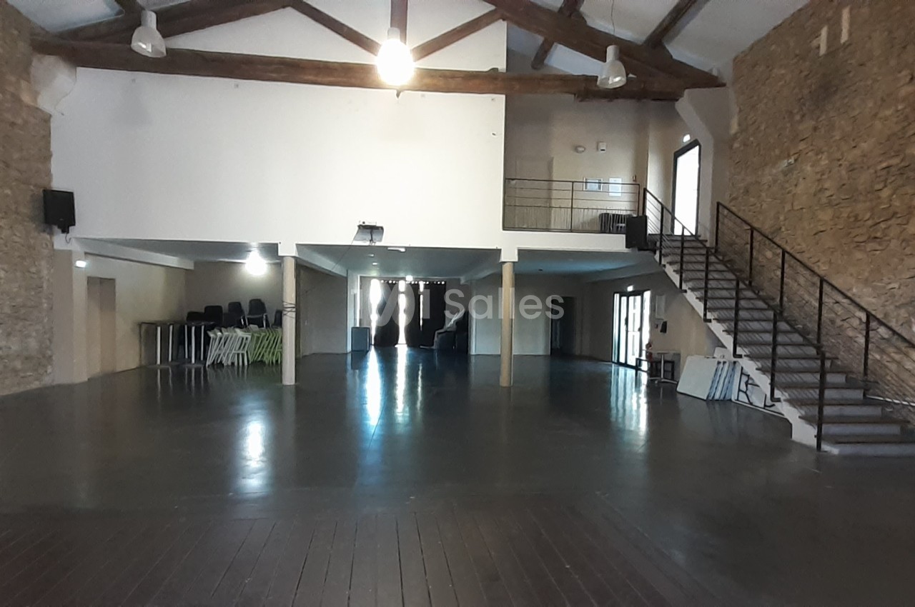 Salle spacieuse avec murs en pierre, plafond à poutres apparentes, escalier métallique et mobilier empilé au fond.