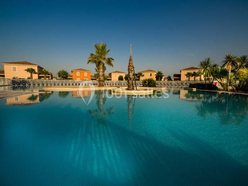 Piscine extérieure entourée de palmiers et de maisons colorées sous un ciel bleu clair.
