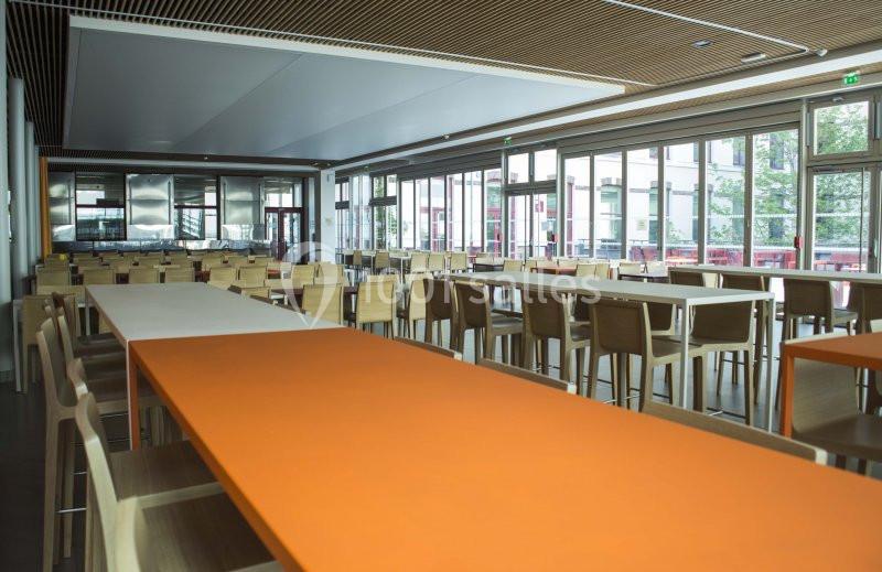 Salle de restauration lumineuse avec de grandes baies vitrées, tables orange et bois, et chaises assorties.