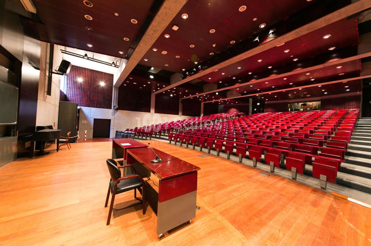 Salle de conférence vide avec rangées de sièges rouges, scène en bois et pupitre équipé d'un micro.