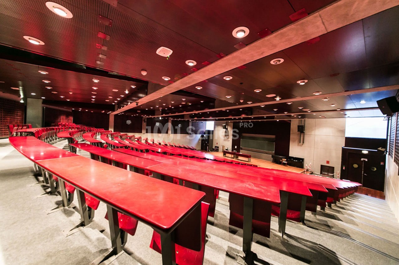 Amphithéâtre vide avec des rangées de tables et sièges rouges, éclairage artificiel et écran au fond.