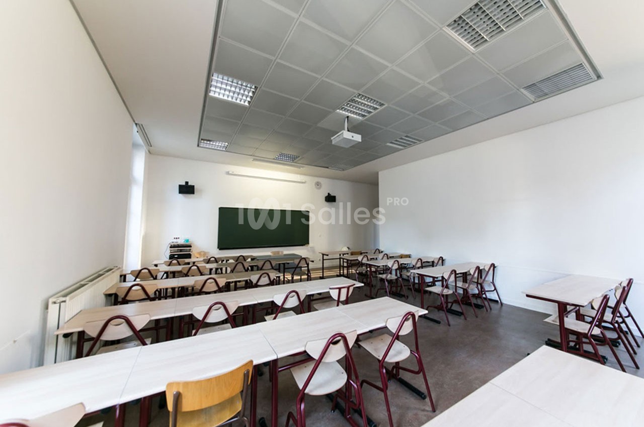 Salle de classe vide avec des tables et des chaises alignées, tableau noir au mur et éclairage au plafond.