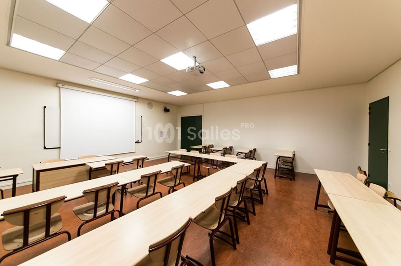 Salle de classe vide avec des rangées de tables et chaises, tableau blanc et projecteur au plafond.