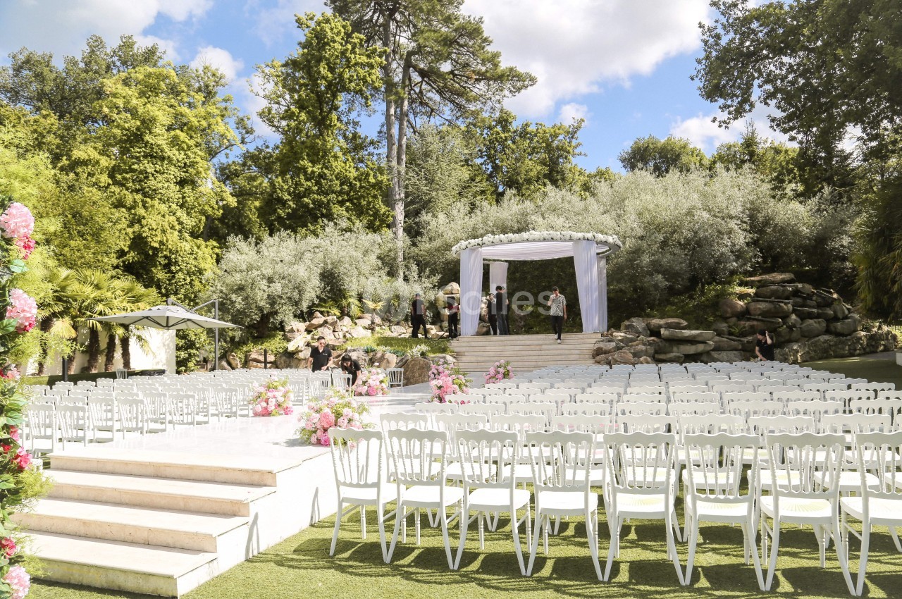 Chaises blanches alignées en extérieur face à une arche décorée pour une cérémonie, entourée de verdure.