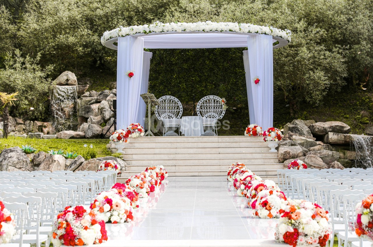 Allée décorée de fleurs menant à une arche blanche ornée pour une cérémonie en extérieur, entourée de verdure.