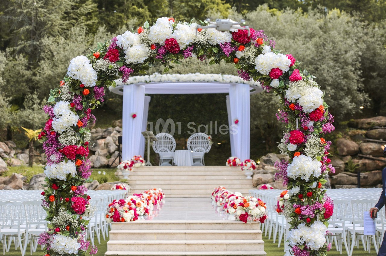 Arche fleurie colorée menant à une cérémonie en plein air avec chaises blanches et décorations florales.