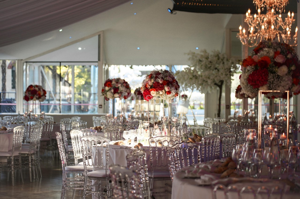 Salle de réception élégante avec tables dressées, chaises transparentes et centres de table fleuris rouges et blancs.