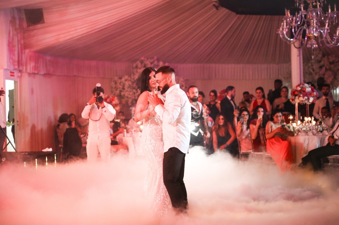 Un couple danse au centre d'une salle de réception décorée, entouré de fumée et d'invités observant la scène.