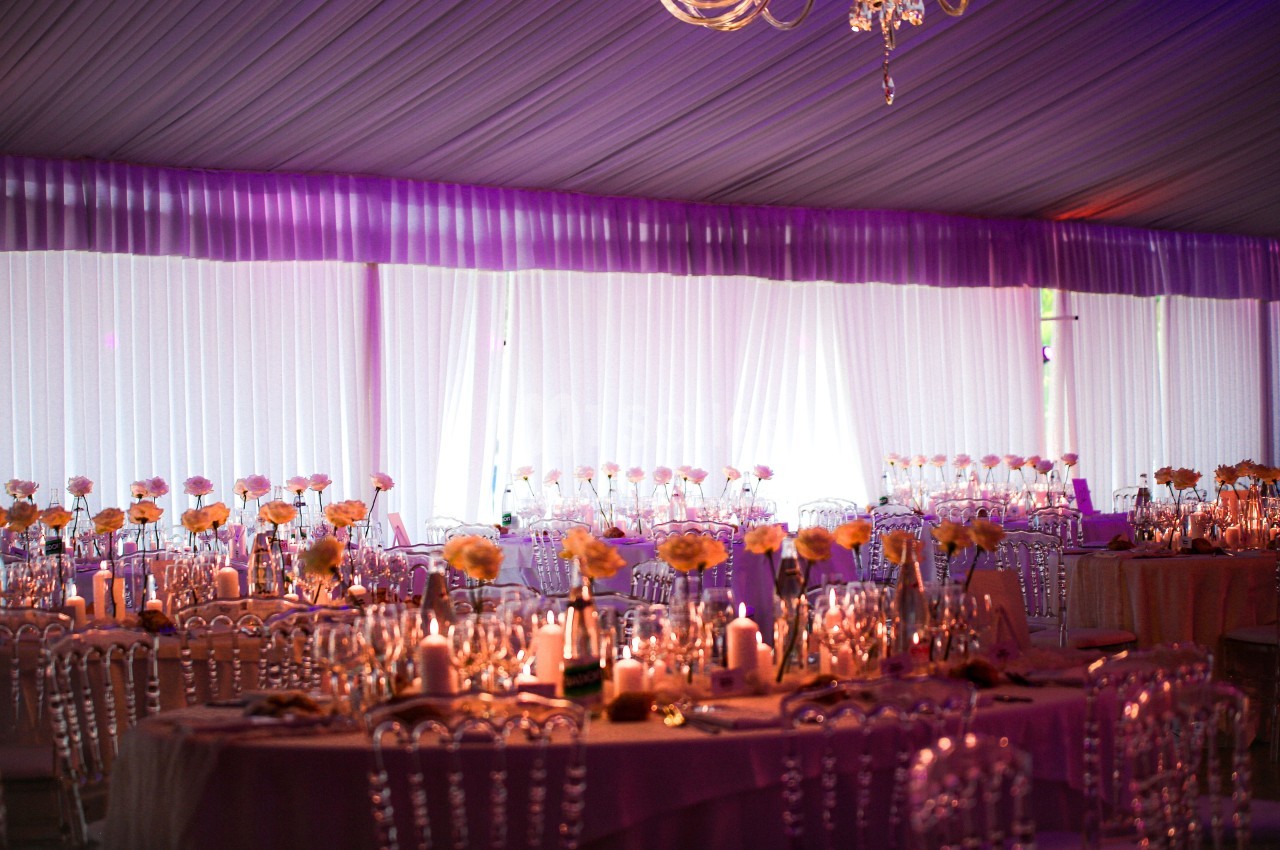 Salle de réception décorée avec des tables rondes, nappes roses, chaises transparentes et centres de table fleuris.