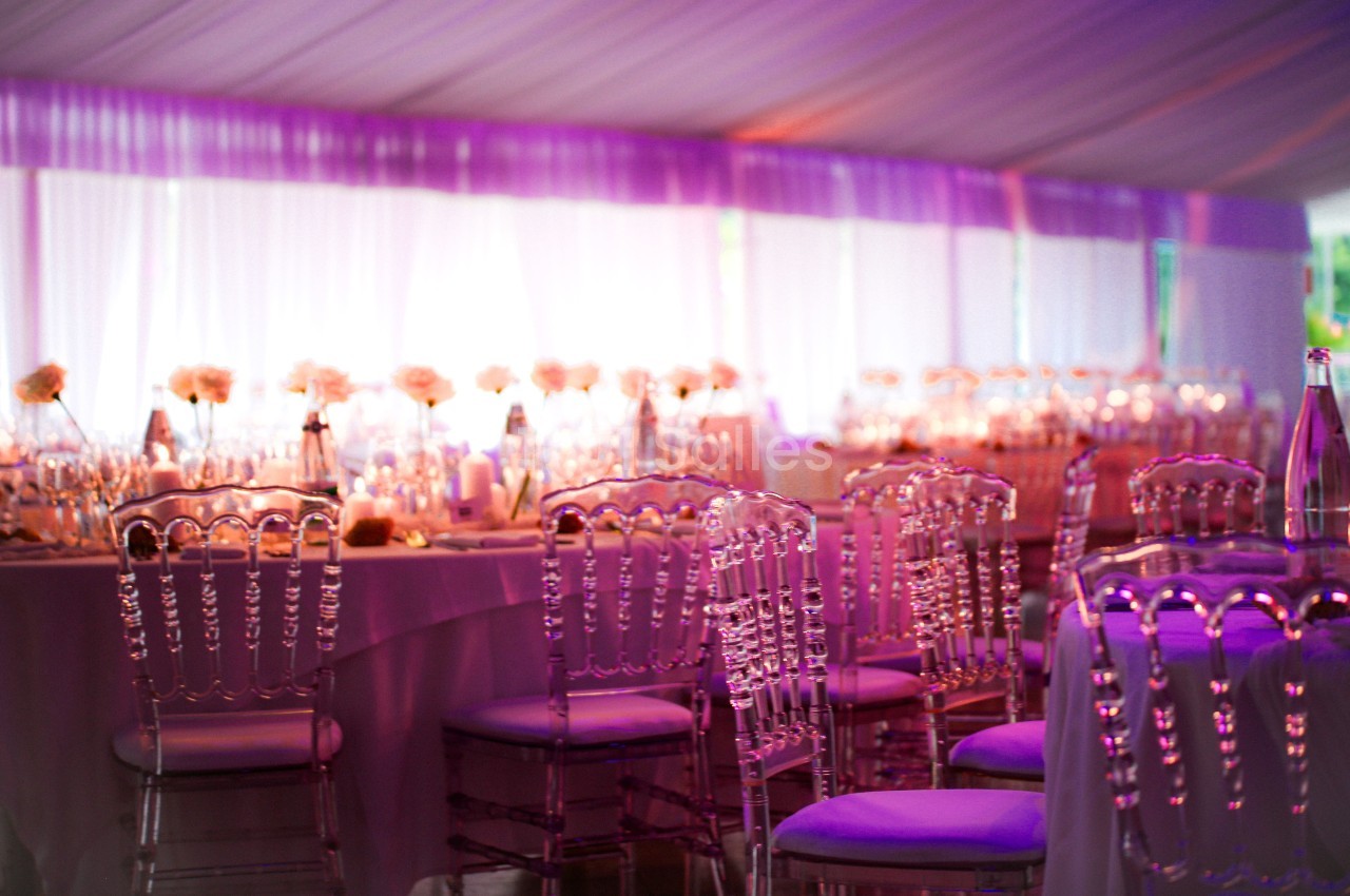 Salle de réception décorée avec des chaises transparentes et des tables ornées de fleurs sous un éclairage violet.