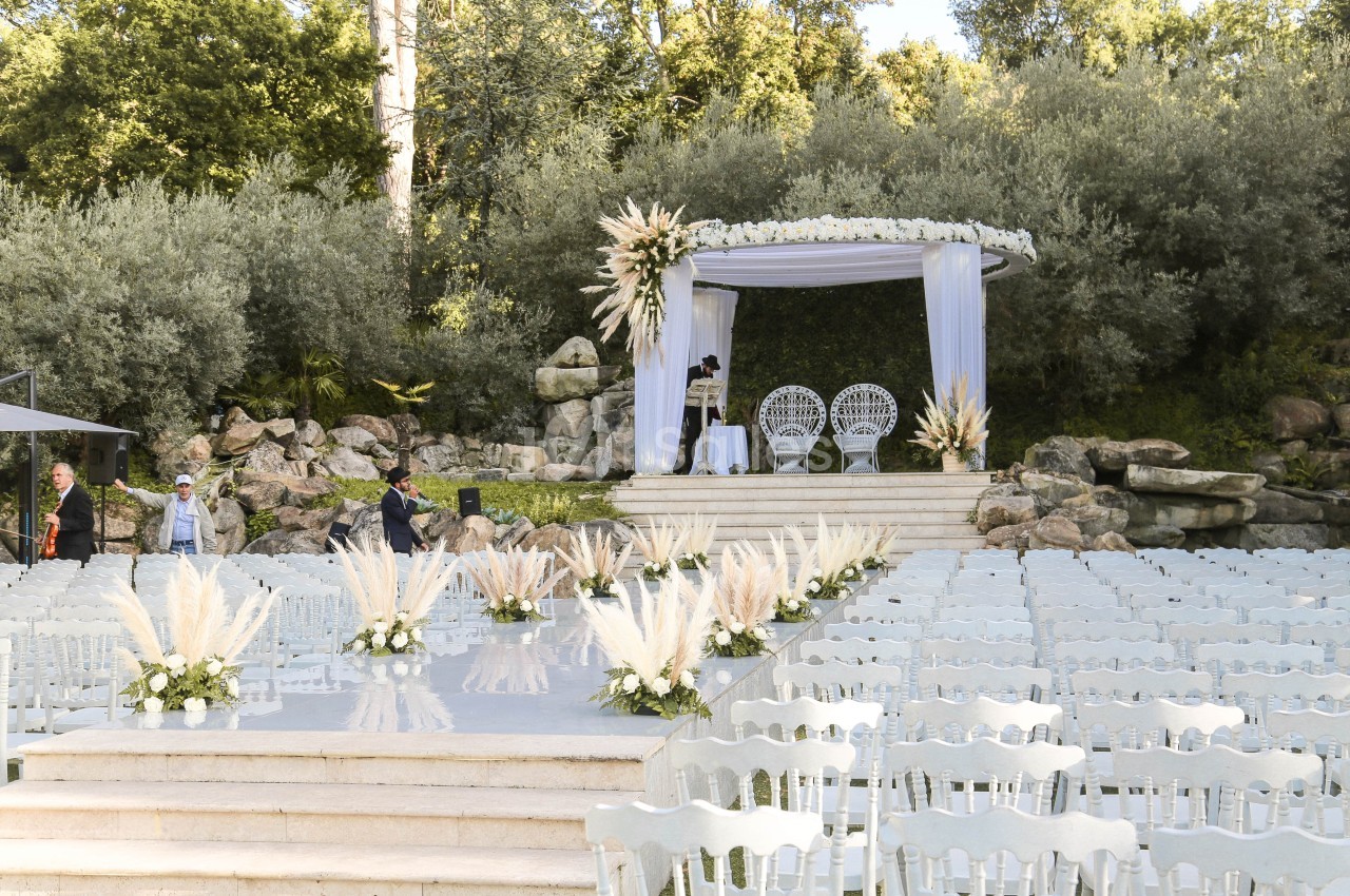 Allée centrale bordée de fleurs menant à une arche de mariage décorée, entourée de chaises blanches en extérieur.