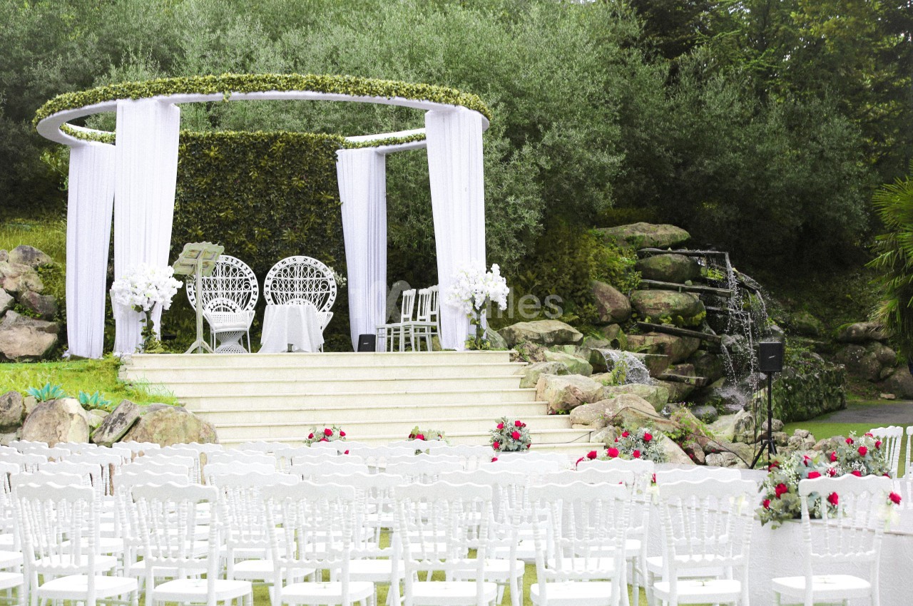 Cérémonie de mariage en extérieur avec arche décorée, chaises blanches alignées et cascade en arrière-plan.