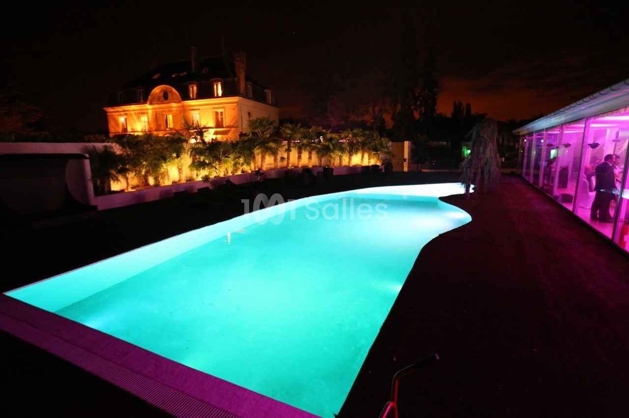 Piscine éclairée de nuit devant une grande maison illuminée, entourée de végétation et d'une terrasse.