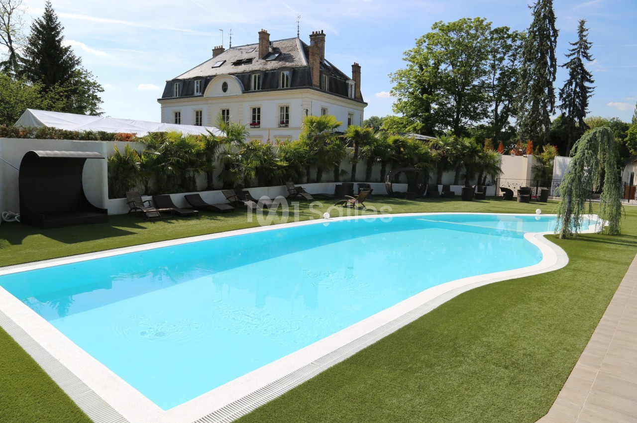 Piscine extérieure entourée de transats, pelouse synthétique et végétation, avec une grande maison en arrière-plan.