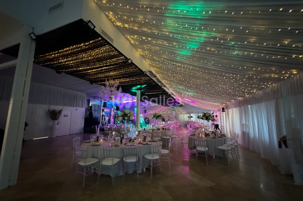 Salle de réception décorée avec des guirlandes lumineuses suspendues, tables rondes dressées et éclairage coloré.