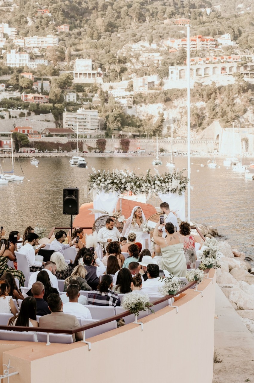 Cérémonie de mariage en plein air au bord de l'eau, avec des invités assis et un paysage urbain en arrière-plan.