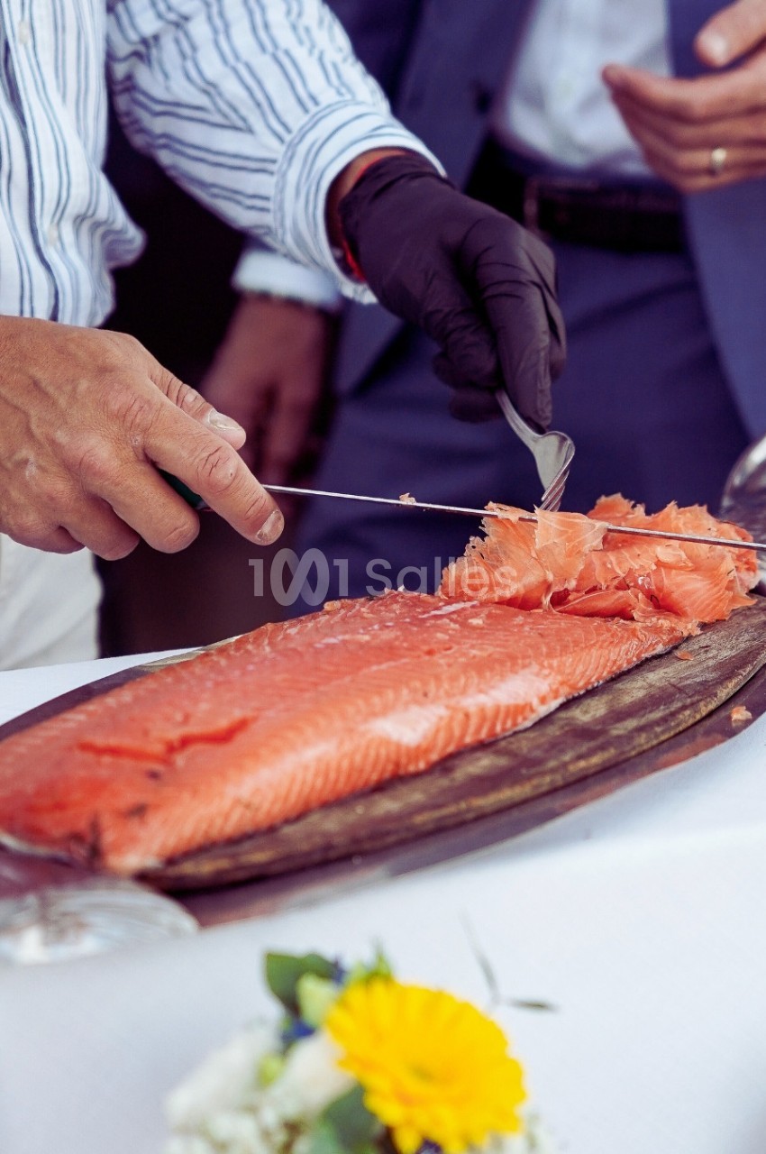 Un homme tranche un filet de saumon frais posé sur une planche en bois, avec une table décorée en arrière-plan.