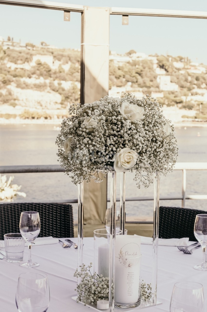 Centre de table floral avec roses blanches et gypsophile, disposé sur une table dressée en extérieur près d'un plan d'eau.
