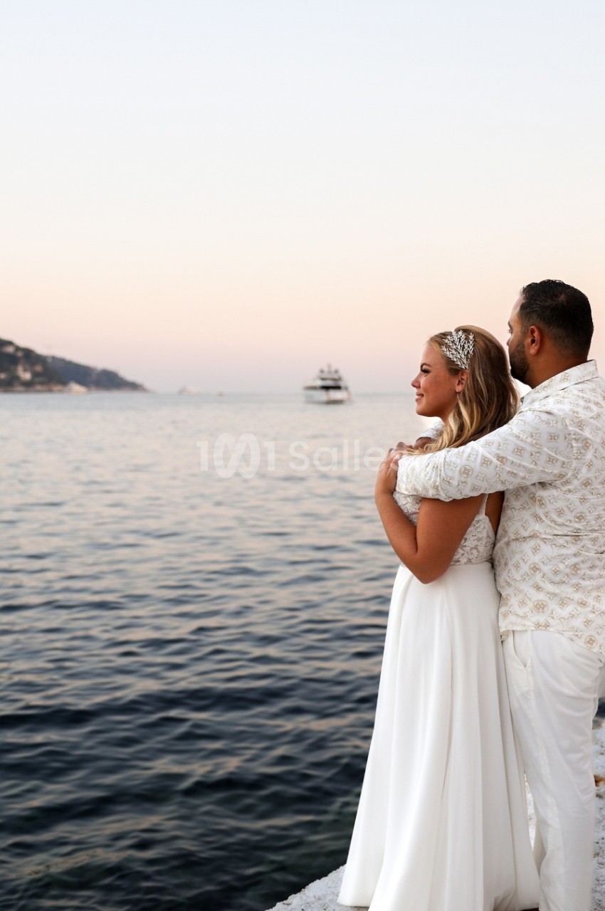 Un couple en tenue élégante regarde la mer au coucher du soleil, avec un bateau visible à l'horizon.