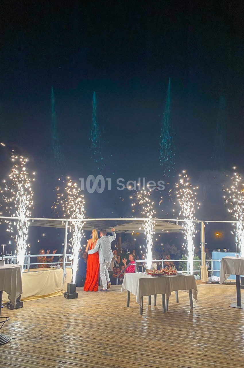 Un couple debout sur une terrasse illuminée par des feux d'artifice, avec des tables dressées en arrière-plan.