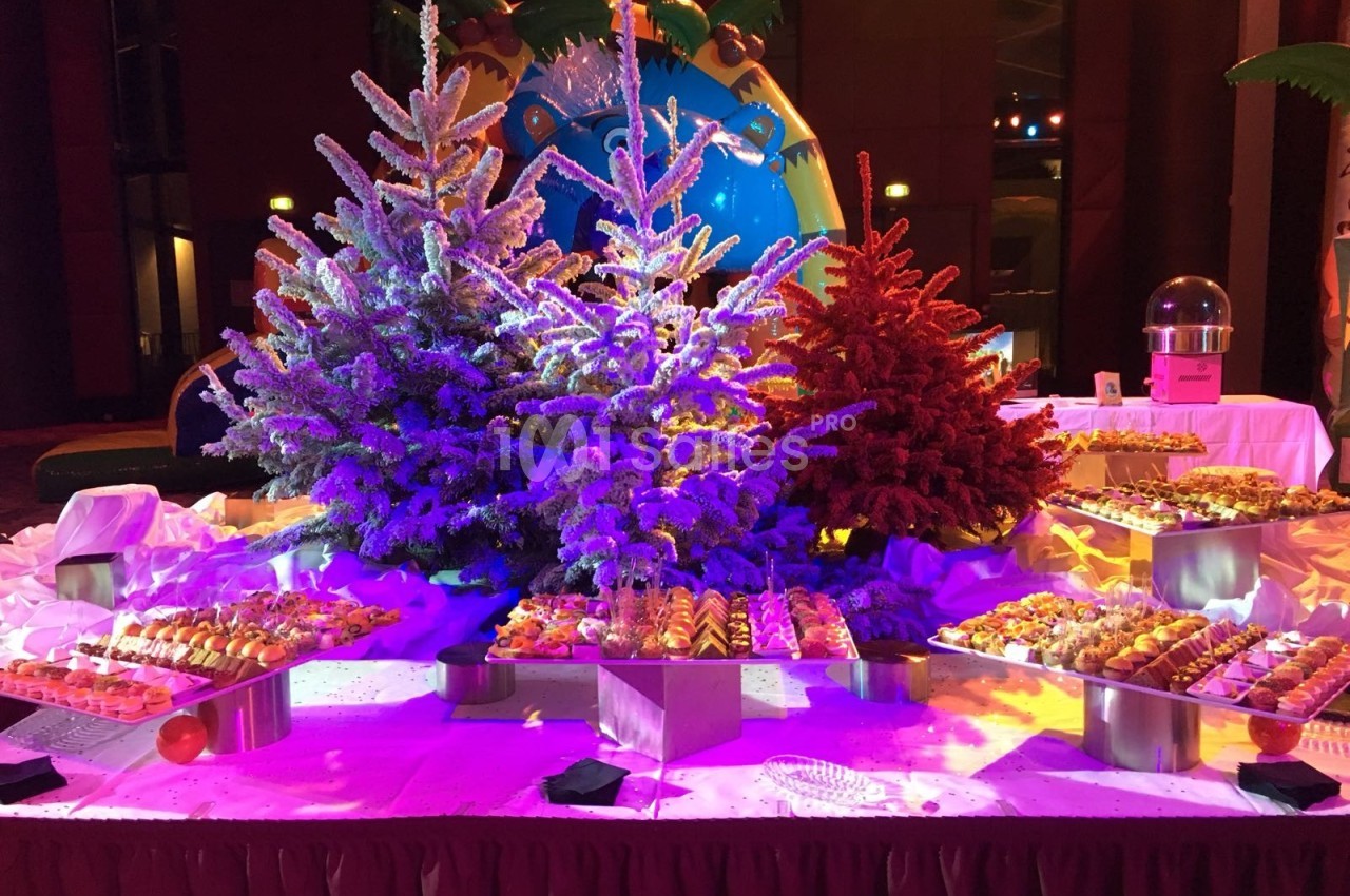 Buffet de desserts présenté devant des sapins décoratifs colorés, éclairés par des lumières vives dans une salle festive.