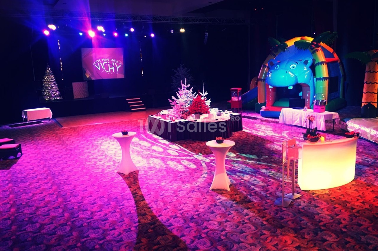 Salle décorée pour un événement festif avec éclairage coloré, tables hautes, sapins et structures gonflables.