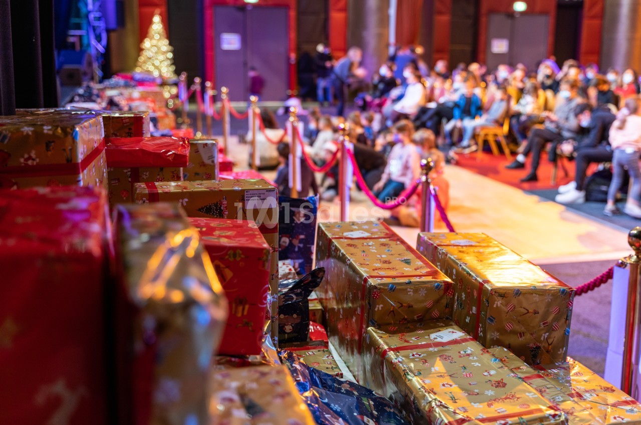 Des cadeaux emballés sont empilés au premier plan, avec un public assis en arrière-plan dans une salle décorée.