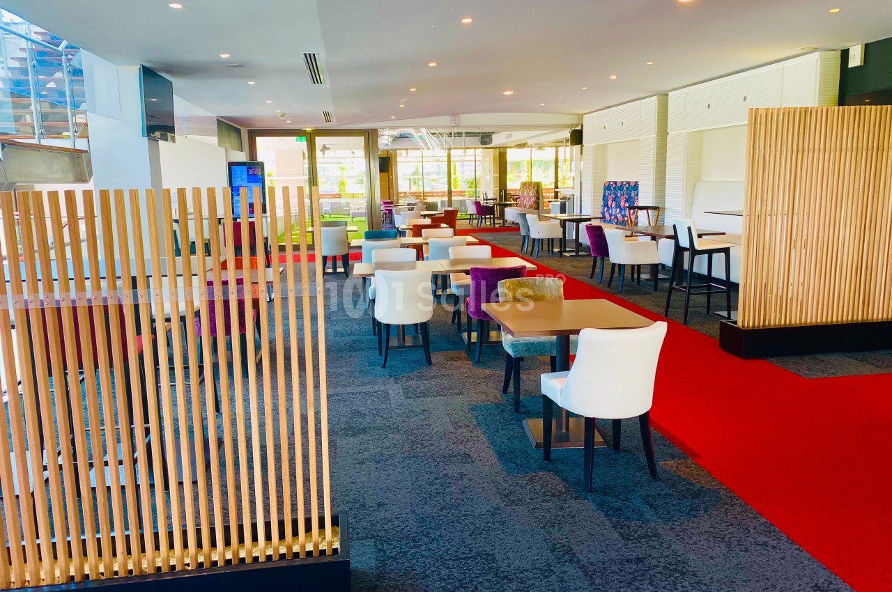 Salle lumineuse avec tables et chaises modernes, sol en moquette, cloison en bois et tapis rouge au centre.