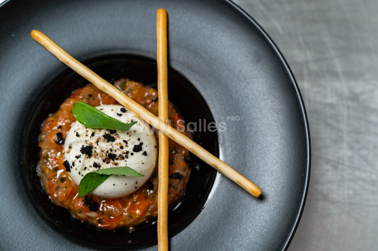 Assiette de burrata sur lit de tartare de tomates, décorée de grissini, basilic et éclats d'olives noires.