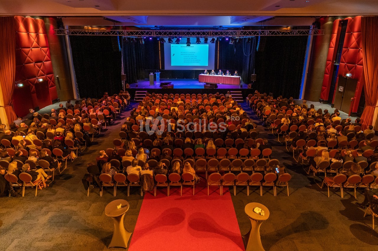 Salle de conférence avec un public nombreux, une scène éclairée et un écran projetant une présentation.