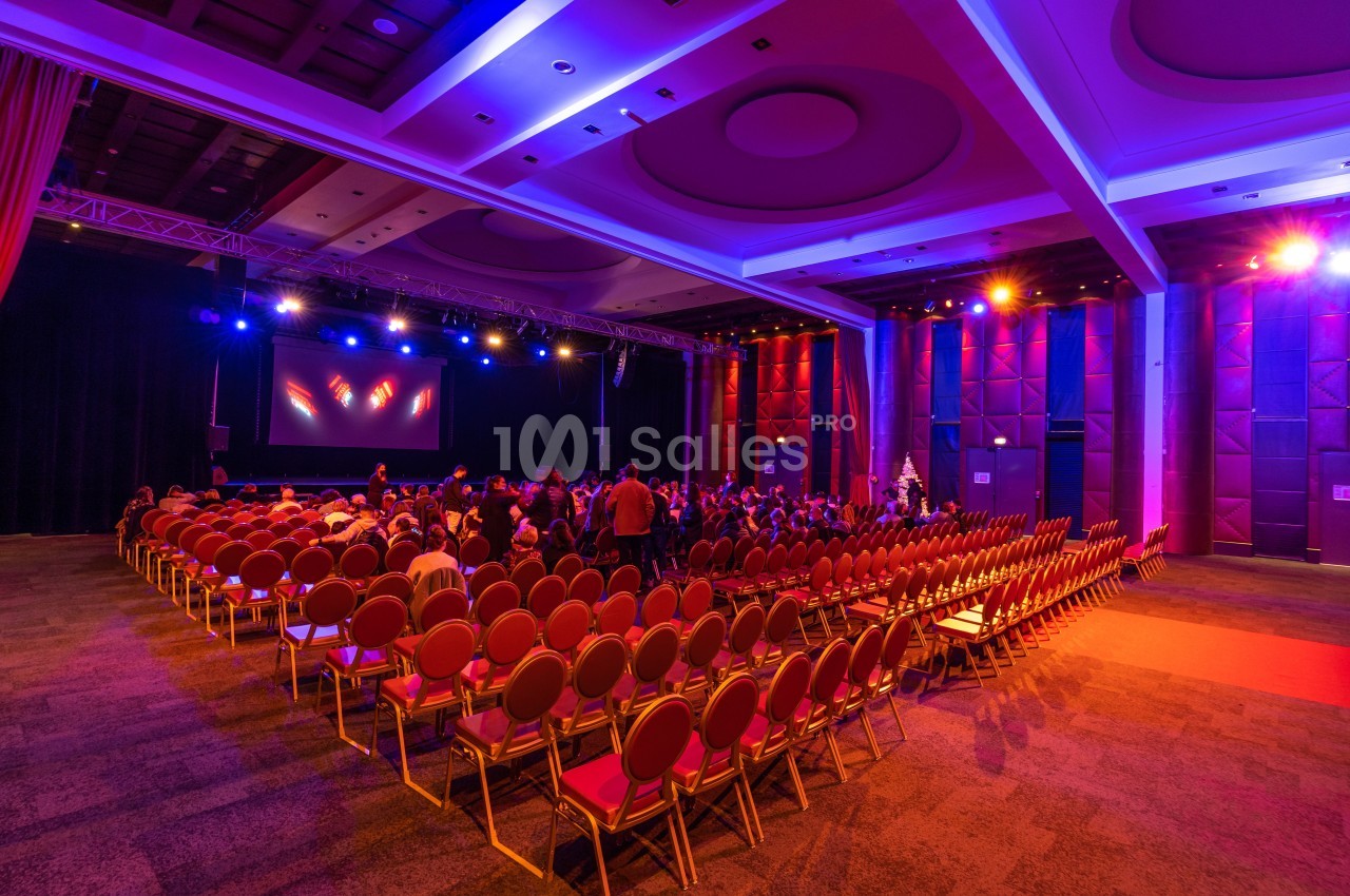 Salle de conférence avec des rangées de chaises rouges, un écran de projection et un éclairage coloré.