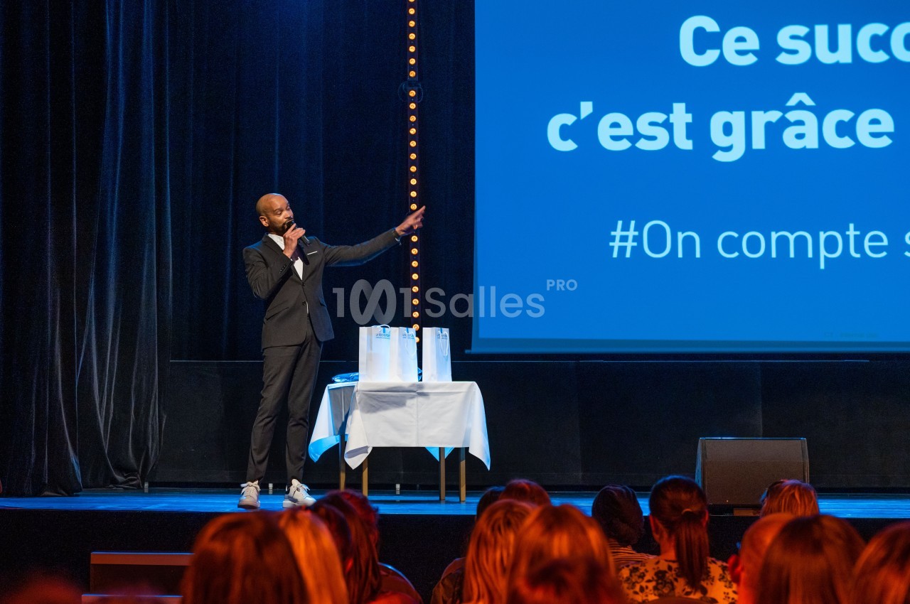 Un homme en costume parle sur scène avec un micro, devant un écran projetant un message et un public assis.