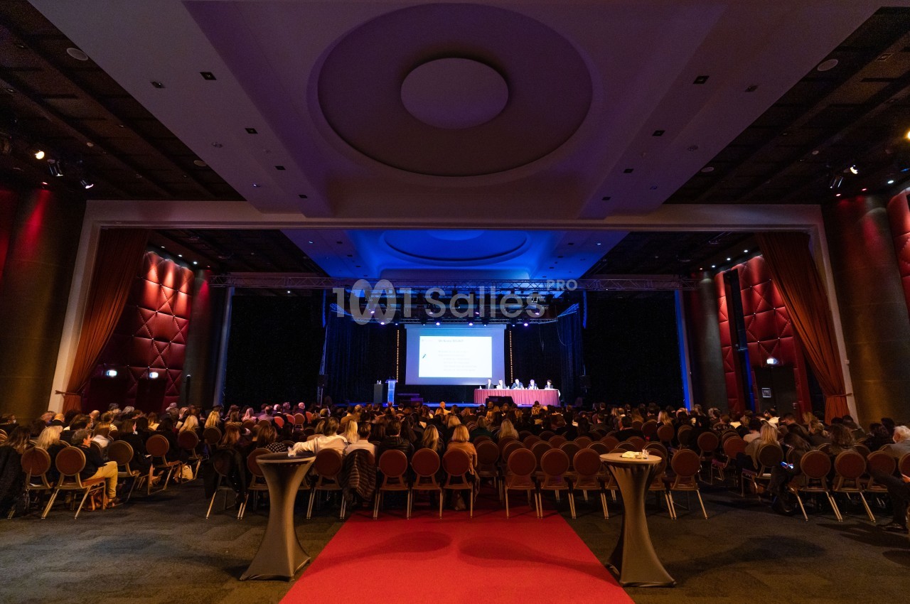 Salle de conférence avec un public assis face à une scène éclairée et un écran de présentation.