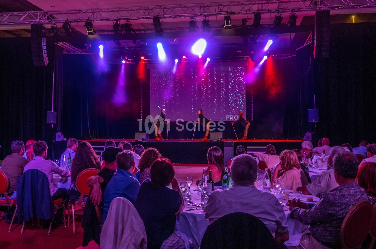 Spectacle sur scène avec trois danseurs sous des lumières colorées, devant un public assis à des tables.