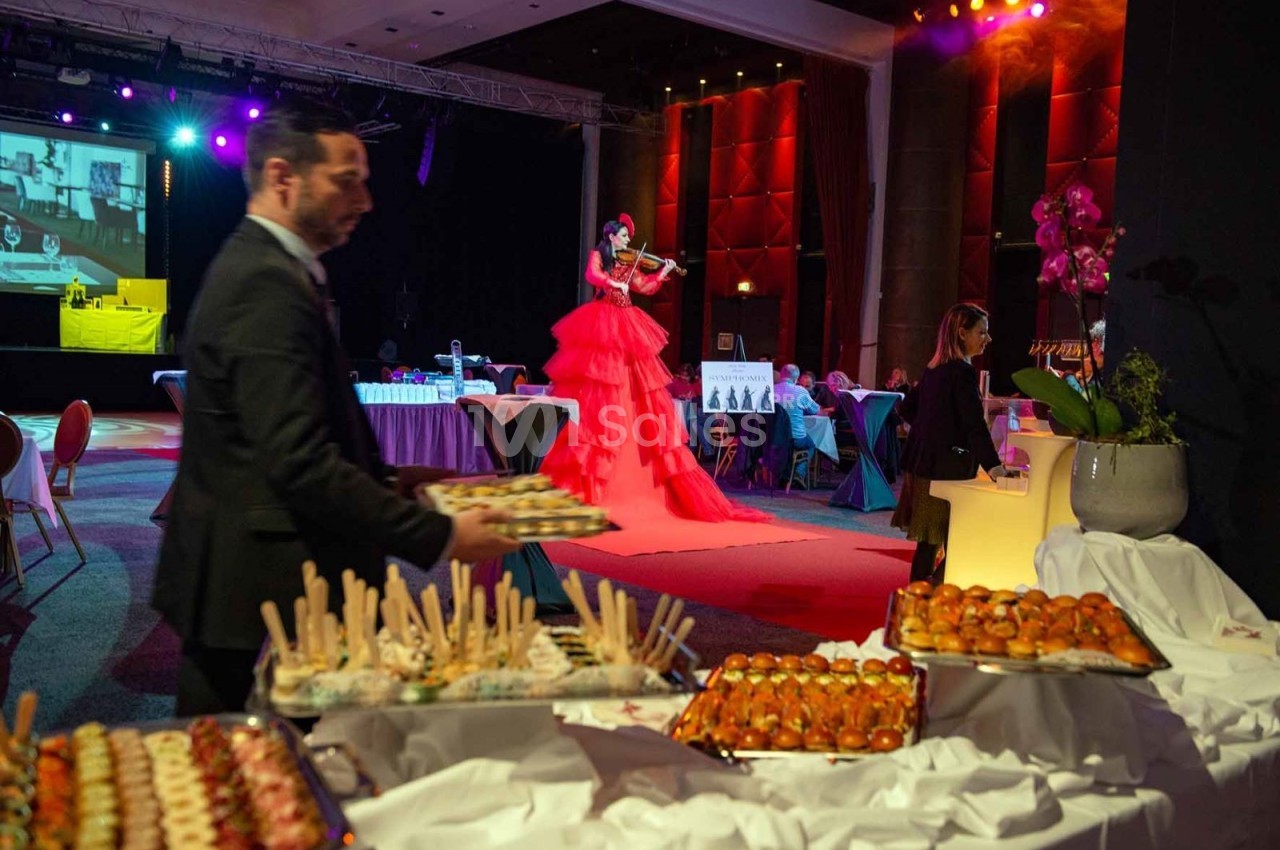 Buffet de plats variés au premier plan, avec une violoniste en robe rouge jouant sur une scène éclairée en arrière-plan.