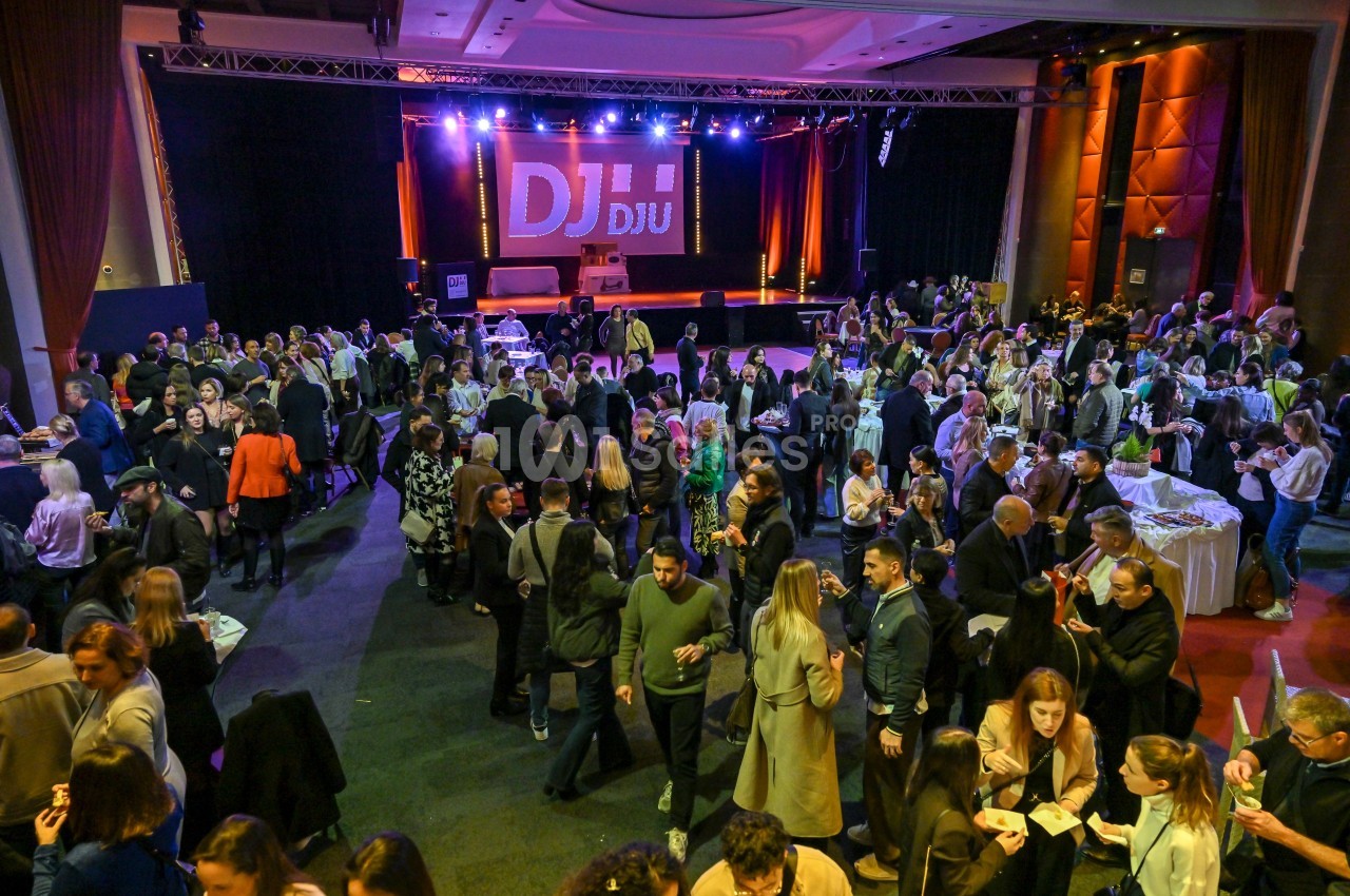 Salle comble avec des personnes debout et discutant lors d'un événement, une scène éclairée avec un DJ en arrière-plan.