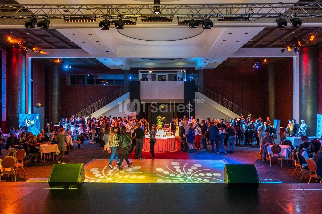 Salle de réception avec des invités debout et assis autour de tables, éclairée par des lumières colorées.